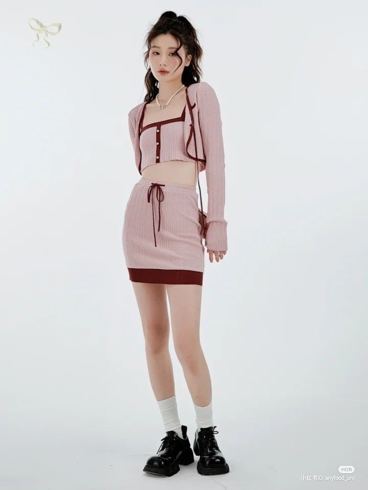 a young woman poses in a studio against a white background. she is wearing a pink threepiece outfit consisting of a cardigan, cropped top, and skirt. the outfit has maroon accents. she is also wearing white socks and black shoes. her hair is pulled back, and she has a neutral expression.