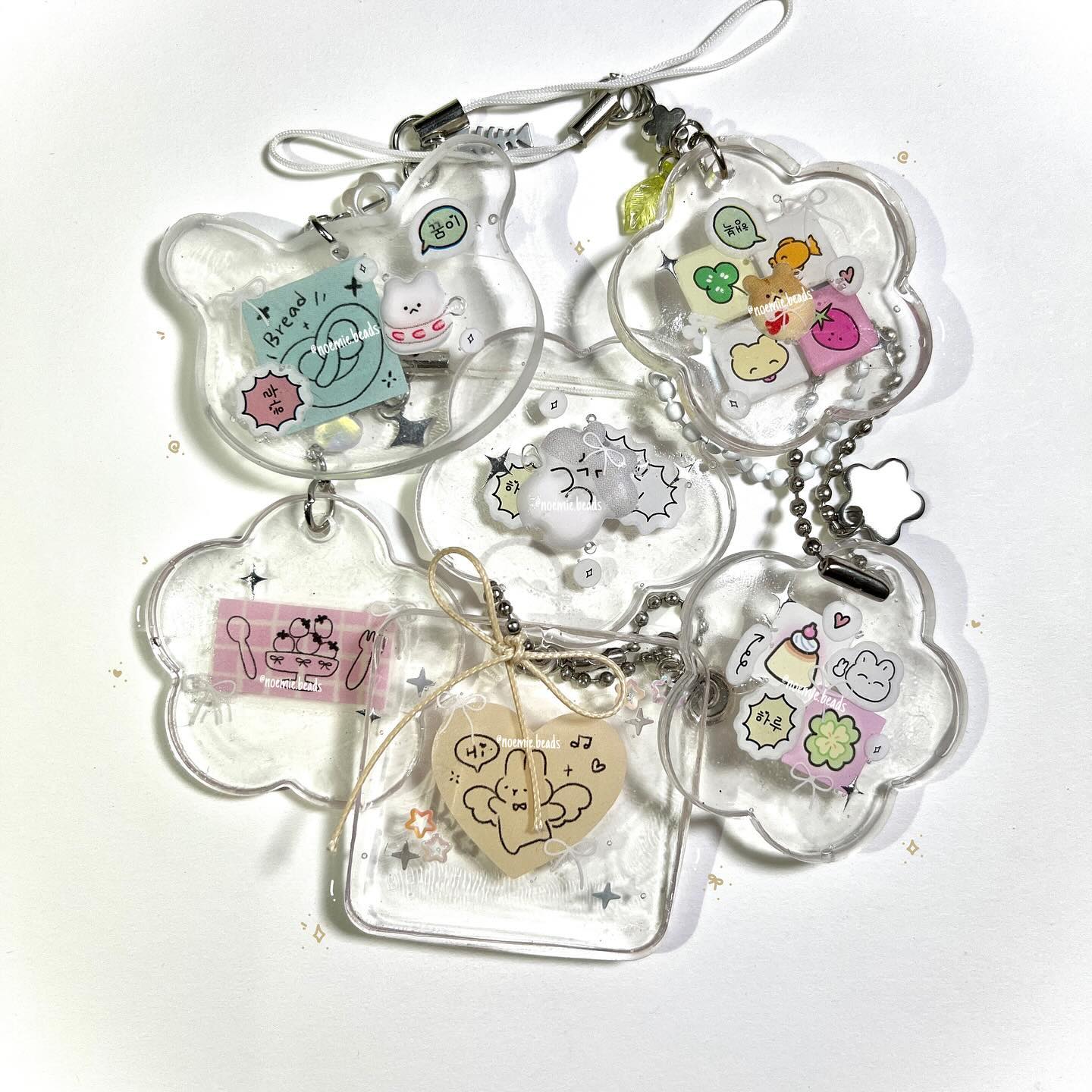 the image shows a collection of acrylic keychains with cute cartoon designs. the charms feature various food and dessert themes, including bread, sweets, and drinks. the keychains are attached to chains and cords. the background is a light surface. the shapes of the acrylic charms are a mixture of clouds, flowers, and hearts. the style is reminiscent of kawaii art.