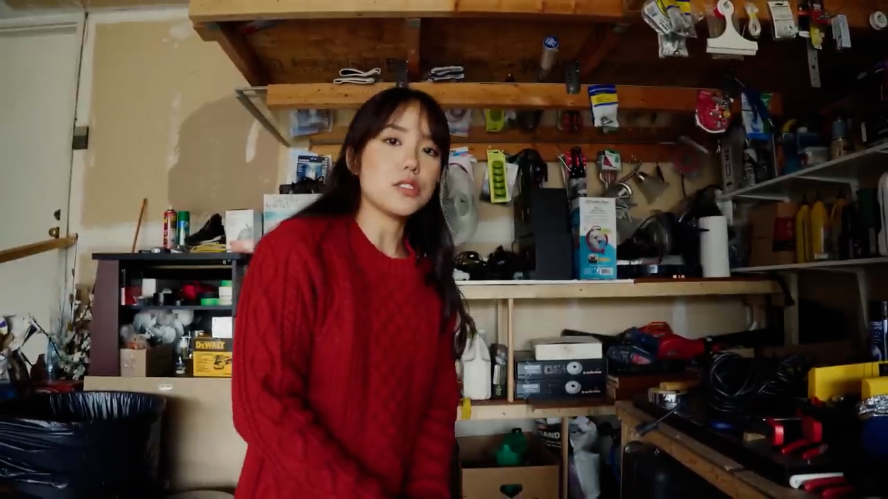 a young woman with long black hair wearing a red sweater is standing in a cluttered garage. she is looking directly at the camera and appears to be talking. the garage has a workbench, shelves filled with tools and supplies, and a window letting in natural light.