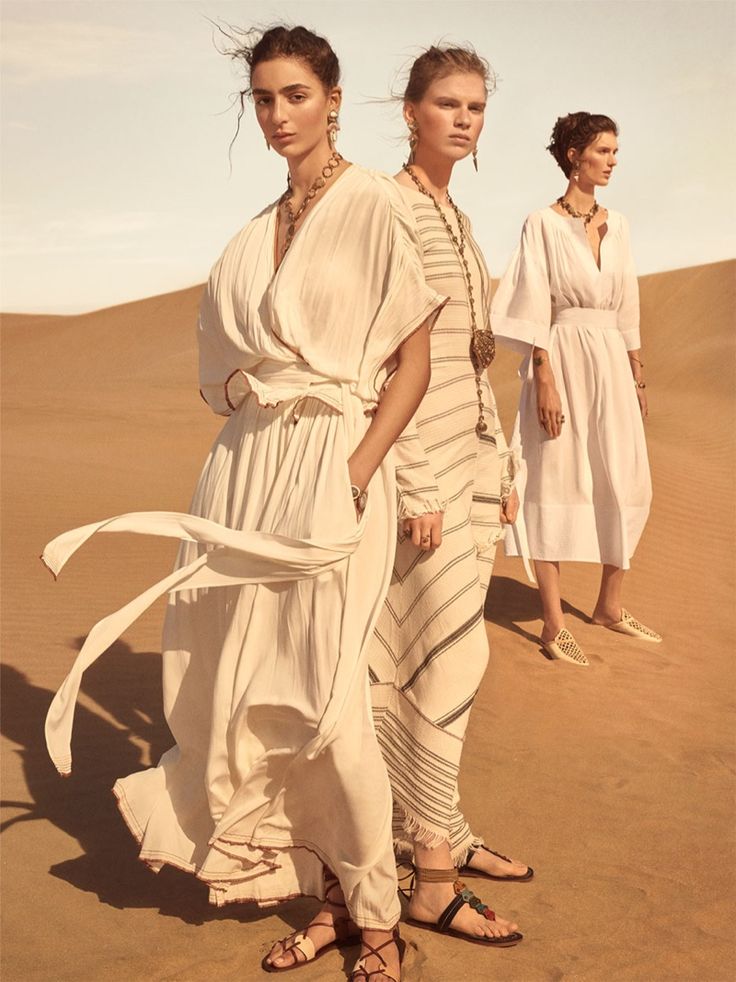 the image is a fashion photograph showcasing three models in long, flowing white and offwhite dresses in a desert setting. the dresses have a bohemian, slightly ethnic feel.  the central model wears a creamcolored pleated maxi dress with a wrap style, while the others wear a striped dress and a simpler white dress. all three are adorned with gold jewelry, including necklaces and earrings.  the models are styled with sandals and their hair is loose. the setting is a sandy desert, bathed in natural light, creating a serene and stylish ambiance. the overall aesthetic is one of relaxed elegance.  the photograph appears to be a promotional image for a clothing brand, possibly zara, considering the clothing style aligns well with the brand's aesthetic.