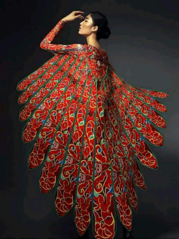 a woman is wearing a long red dress with intricate green designs. the dress is flowing and has a large, dramatic, winglike design. the woman is posing elegantly with her head turned to the side. the background is a dark grey. the dress appears to be handmade and extremely detailed.