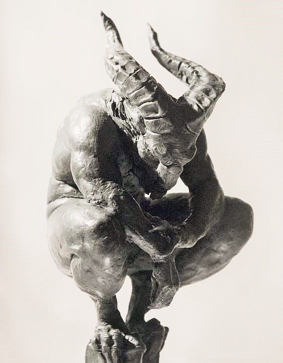 a bronze or clay sculpture of a crouching, muscular, horned figure with its head down. the figure appears demonic, with large, textured horns. it is mounted on a rough, uneven base. the artwork is monochromatic.