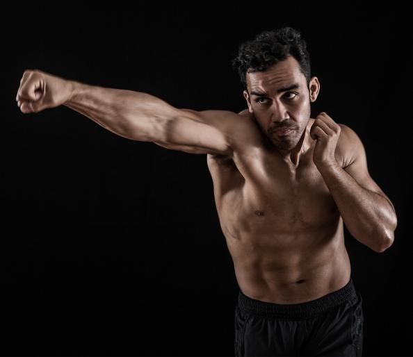 a muscular man in black shorts is punching with his right hand towards the camera. he is posing in a studio with a black background. he is looking off to the side and has a determined expression on his face. he is an athlete or bodybuilder with a toned physique and a sixpack.