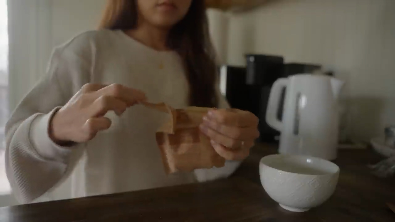 a woman stands in a kitchen and prepares a cup of tea. she holds a brown tea bag in her hand. on the counter in front of her are a white kettle and a white teacup. the scene is illuminated by soft indoor lighting, creating a cozy atmosphere.