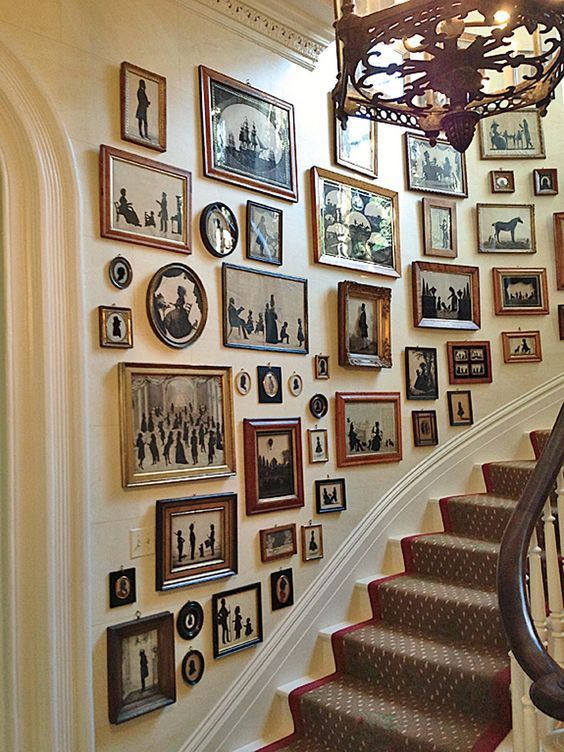 this image shows a staircase with a gallery wall filled with framed pictures, mostly silhouettes. the staircase has a patterned carpet with a red border, and a dark wood banister. the gallery wall features a variety of frames and sizes, creating a visually interesting display. a chandelier hangs above the staircase, adding to the elegance of the space.