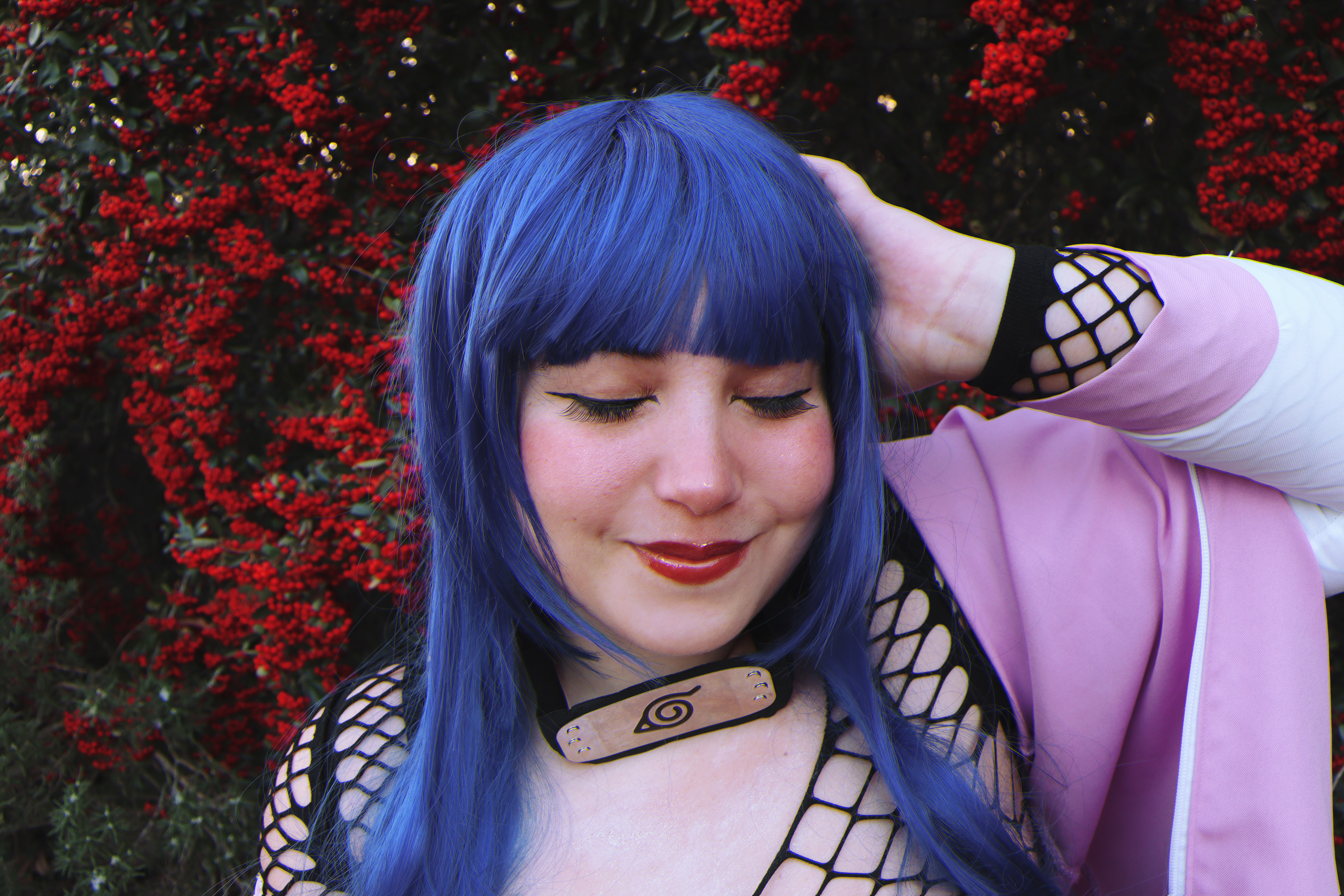 a woman cosplaying as hinata hyuga from naruto, wearing a blue wig, fishnet top, and pink kimono. she has on makeup with eyeliner and red lipstick, and is wearing a headband. she is smiling with her eyes closed in front of a backdrop of red berries.