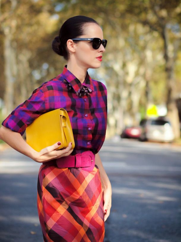 the image shows a stylish woman wearing a vibrant autumnal outfit.  she's posing outdoors on a city street. her outfit consists of a purple, red, and orange plaid shirt worn over a similarly patterned skirt.  she is accessorized with a bright yellow shoulder bag, a fuchsia belt, and dark sunglasses. her hair is pulled back in a sleek updo.  the overall feel is trendy and modern, with a focus on bold colors and patterns. the background is blurred, drawing attention to the woman and her outfit.