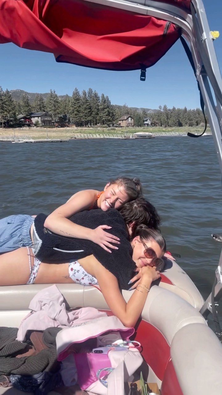 three women hugging on a boat on a lake. there are mountains and trees in the background.