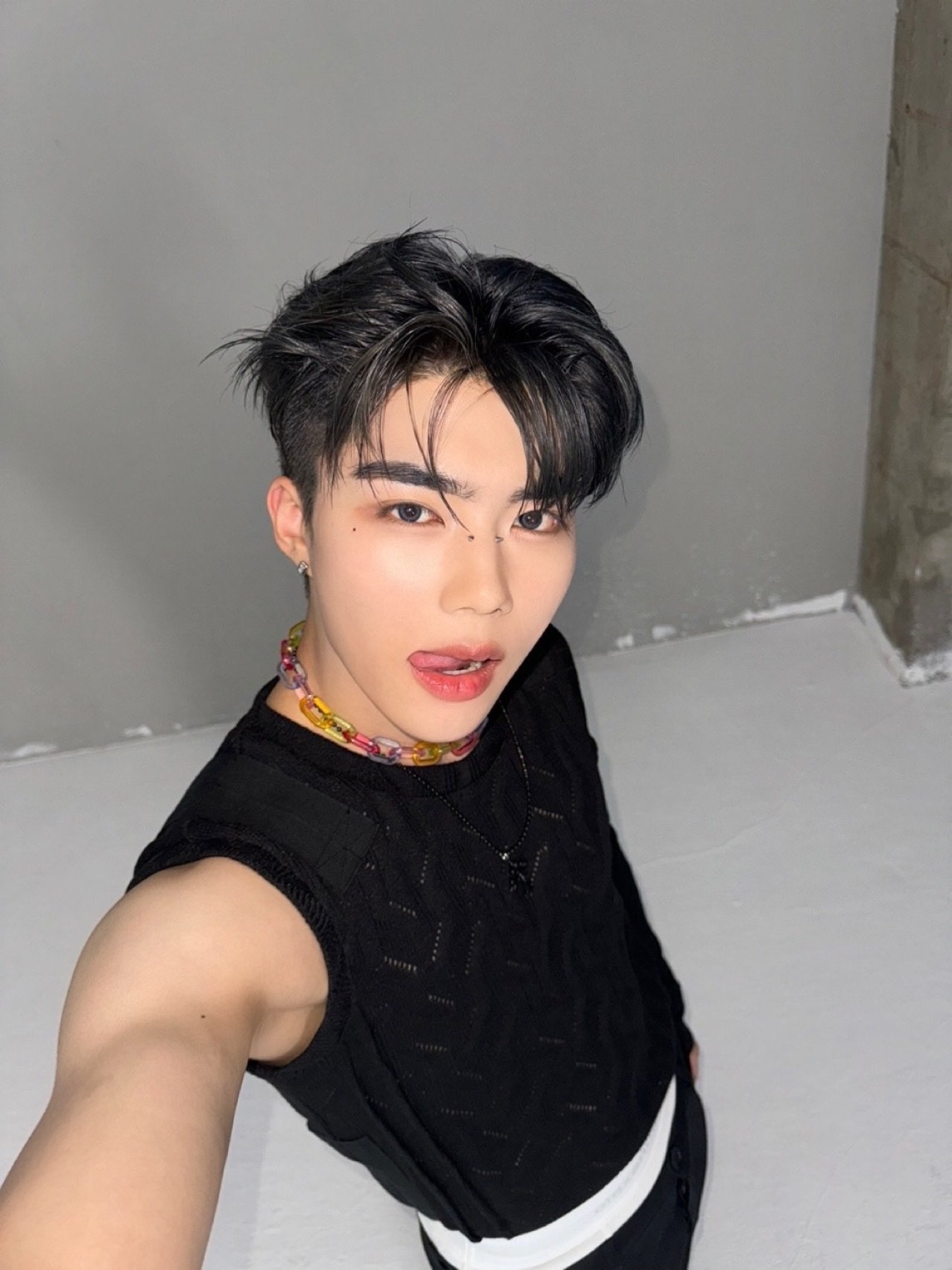 a young asian man takes a mirror selfie. he is wearing a black tank top, black pants with a white stripe, and a colorful beaded necklace. his hair is styled in a trendy cut, and he has a playful expression on his face, sticking out his tongue slightly. the background is a simple gray wall, and the lighting is soft, creating a flattering portrait.