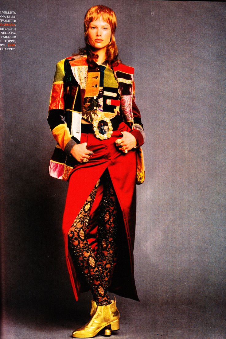 a woman in a fashion editorial stands against a plain background. she wears a patchwork velvet jacket, a red slit dress, lace leggings, and gold ankle boots. her hands are in her pockets and she has a serious expression.