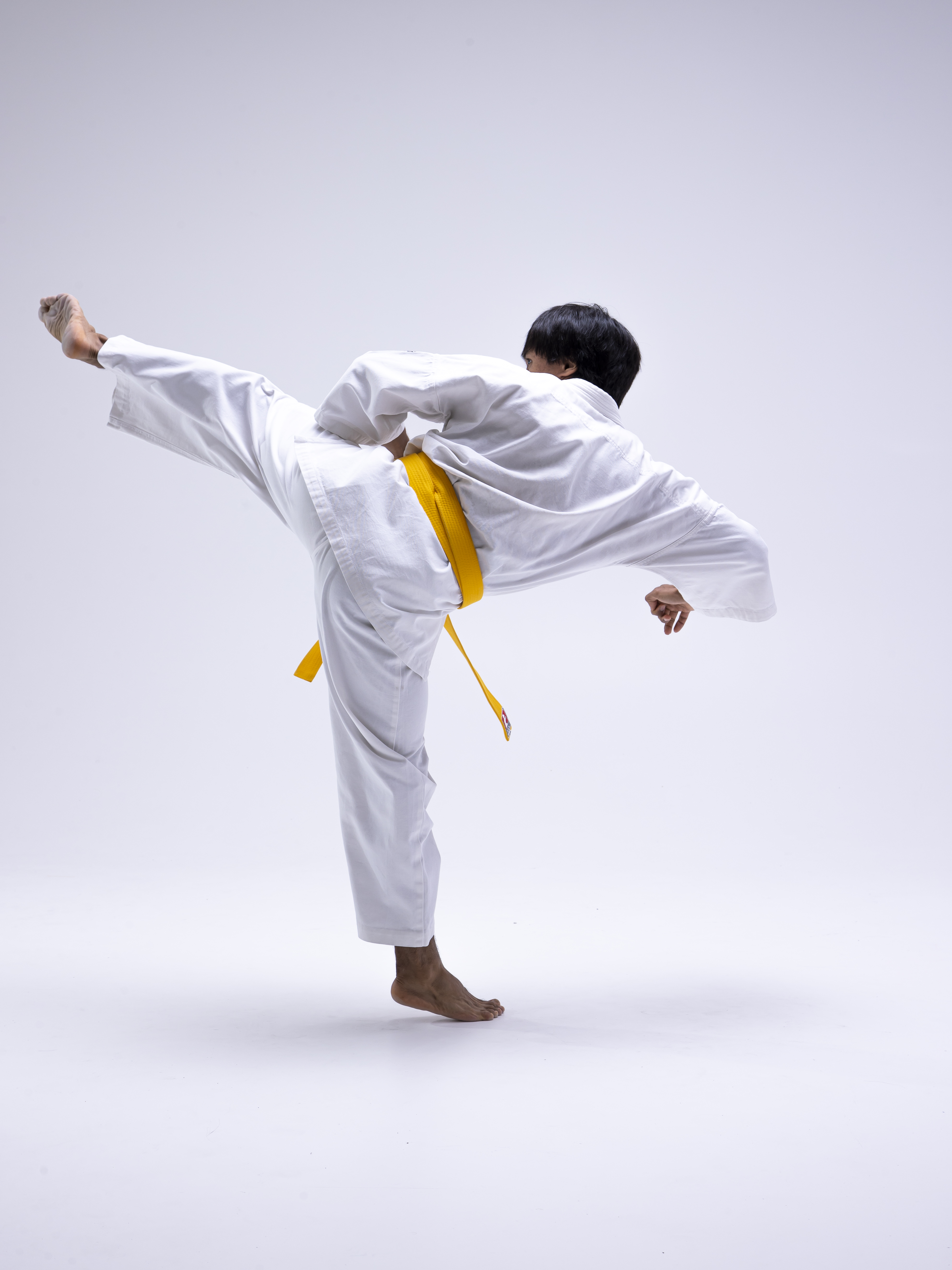 a person wearing a white karate gi with a yellow belt is performing a side kick. they are barefoot and positioned against a plain white backdrop. the image is a full body studio shot capturing the dynamic pose.