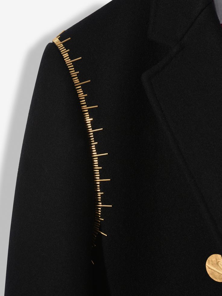 loseup view of a black coat or blazer featuring a unique gold metallic embellishment along the shoulder seam. the embellishment resembles a series of small, evenly spaced gold studs or pins creating a linear pattern. the coat itself appears to be made of a thick, possibly woolen fabric. the overall style is elegant and luxurious.  the image is sharply focused, showcasing the texture and detail of both the fabric and the gold embellishment.  a small portion of a gold button is visible near the bottom right corner.