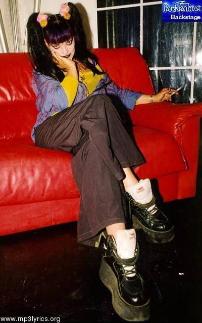 a young goth woman wearing black platform shoes and a  black and yellow grunge outfit sits on a red couch. she is looking at the camera.