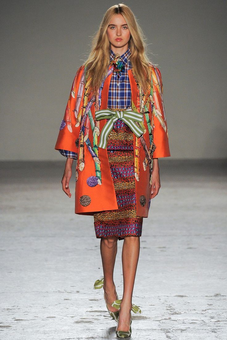 a full shot of a fashion model walking down a runway. she is wearing a vibrant orange coat with a whimsical printed pattern of what appears to be stalks of sugarcane or similar plants. underneath, she sports a colorful, patterned pencil skirt. a plaid shirt is visible at the collar and cuffs.  she's accessorized with a light green sashlike belt and eyecatching shoes with bows. her long blonde hair is styled simply, and her expression is neutral and professional. the background is a neutraltoned runway. the overall aesthetic is bold and artistic, highlighting a unique and eyecatching design.