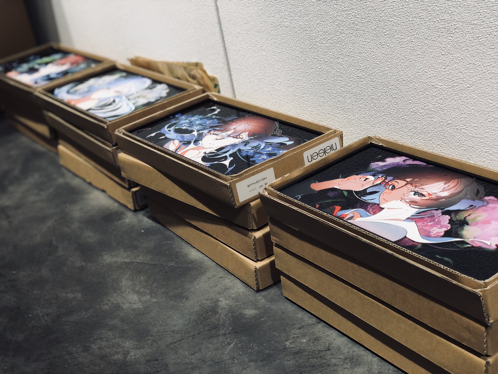 stacks of artwork featuring animestyle characters are neatly arranged on the floor, likely prepared for shipping or storage. the boxes are uniformly brown and labeled, hinting at a professional art distribution setting. the vibrant colors and intricate details of the artwork suggest highquality prints.