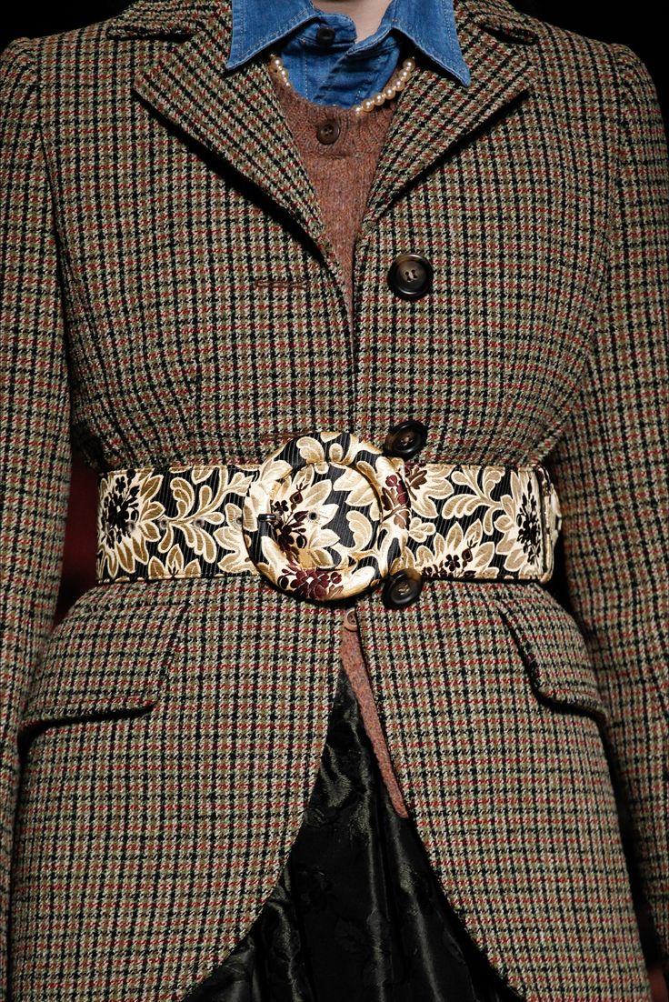 loseup of a woman wearing a brown and beige tweed jacket with a patterned floral belt featuring a large, ornate buckle.  the jacket has a houndstooth pattern. the woman is also wearing a denim shirt, a brown sweater, and a pearl necklace, all visible underneath the jacket. the overall style is vintageinspired and autumnal in its color palette. the image focuses on the texture and detail of the clothing and accessories.