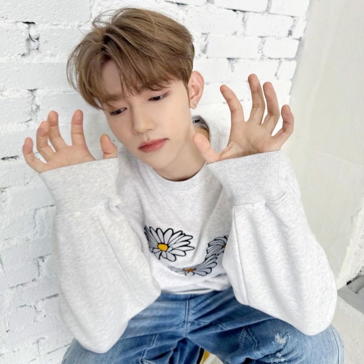 a young man poses for a photo. he's wearing a white sweatshirt with a daisy design on it and ripped jeans. he has his hands up by his face in a playful pose. the background is a white brick wall. the lighting is soft and natural.
