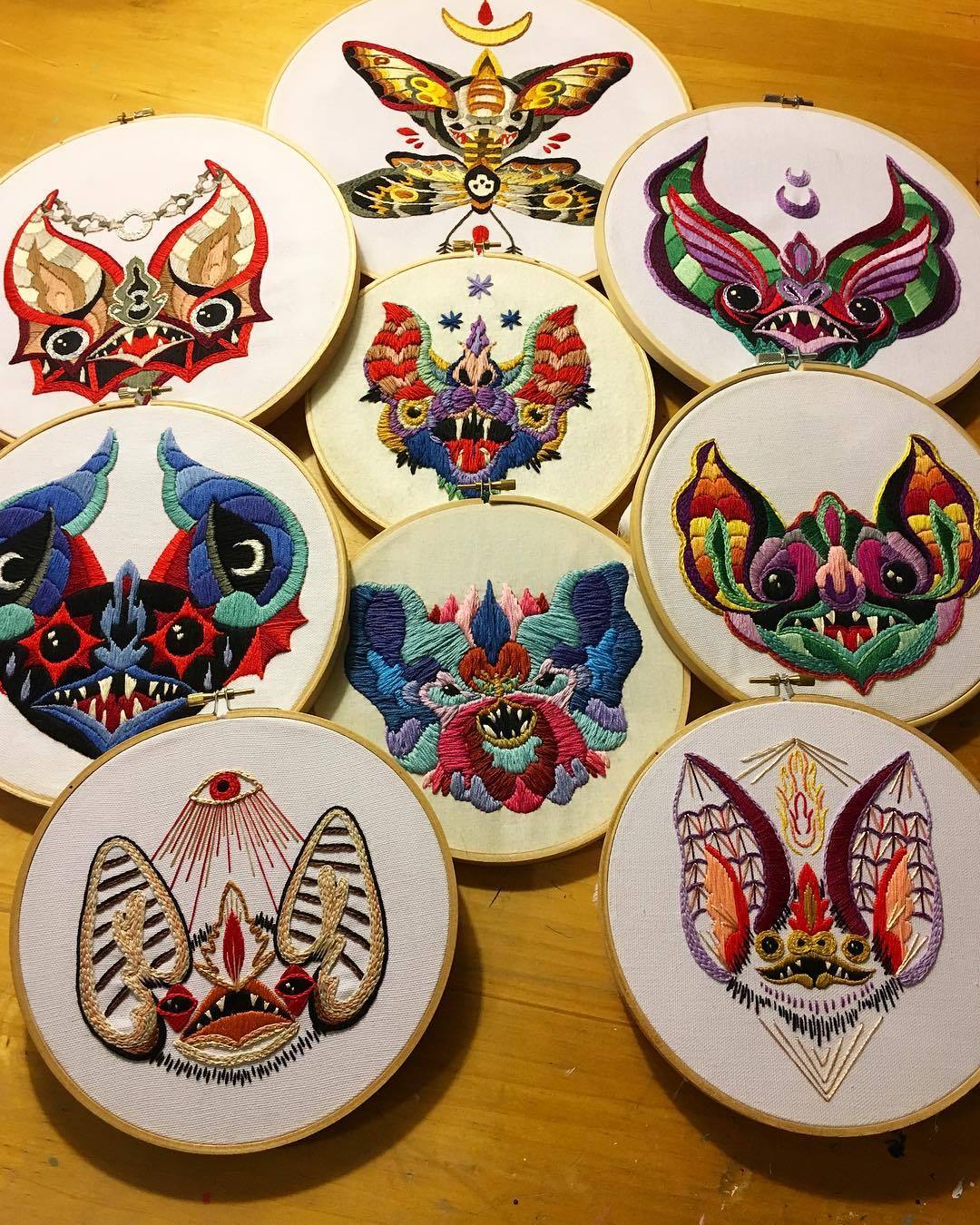the image showcases a collection of ten vibrant and intricately embroidered pieces of art. each piece is circular, enclosed in an embroidery hoop, and features a unique design centered around bats or a moth. the bats are depicted in various styles, from cartoony and whimsical to more grotesque and gothic. the embroidery is incredibly detailed, with a rich variety of colors and textures used to create depth and visual interest. the background is a light wooden surface, providing a neutral backdrop to highlight the colorful artwork. the overall aesthetic is a mix of folk art, whimsical design, and darker gothic elements, creating a captivating and eyecatching display.