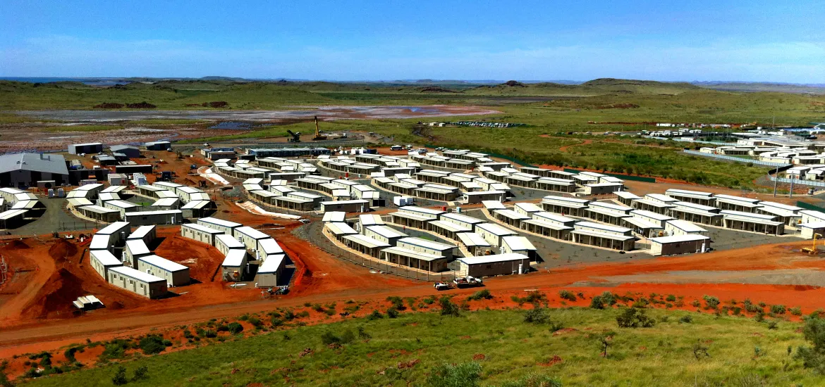 an aerial view of a remote mining town in the australian outback. the town is made up of a large number of prefabricated buildings, most likely housing workers at the mine. the town is surrounded by red dirt and desert landscape. the image is taken from a drone and shows the vastness of the australian outback and the isolation of the mining town.