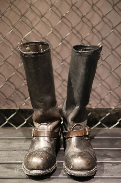 a pair of brown leather motorcycle boots with black steel toes are placed on a wooden floor. the boots are worn and distressed, giving them a rugged and tough look. the boots have a vintage feel and are perfect for casual street wear.