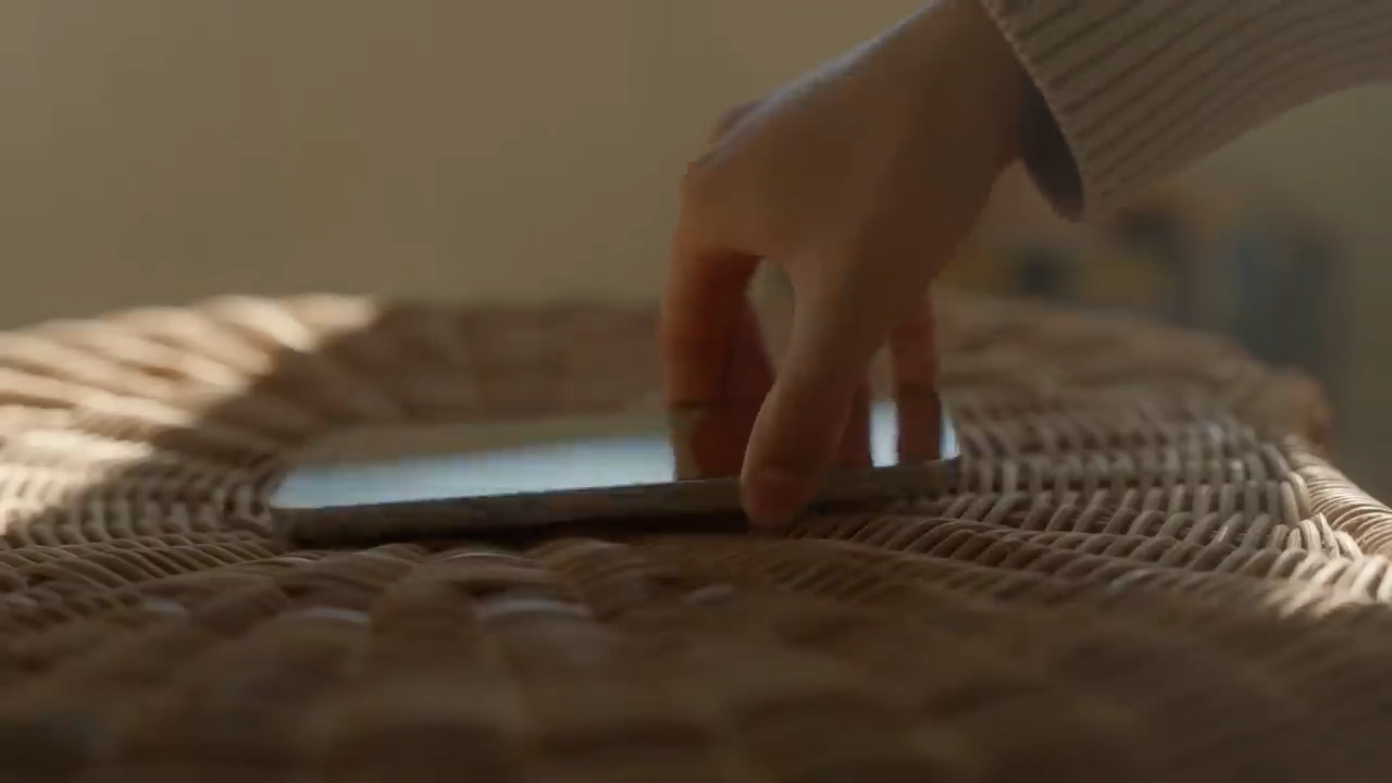 a hand is placing a smartphone on a rattan table with sunlight shining through a window.