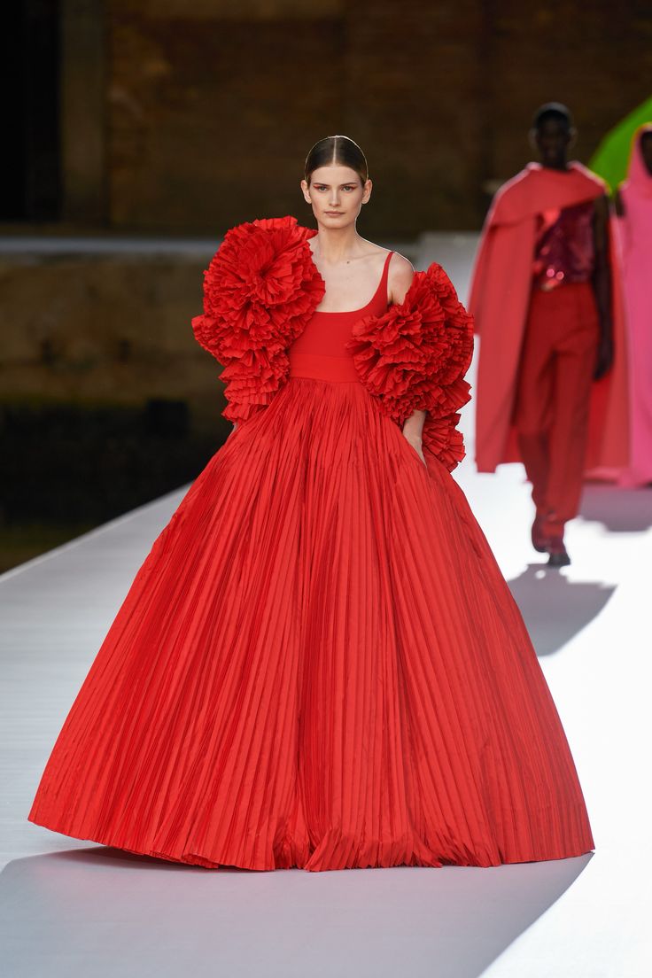 a woman in a red gown with ruffled sleeves walks down a runway. her gown is pleated and falls to her feet, creating a dramatic silhouette. the woman is smiling and looks confident as she walks, showing off the elegant design of her outfit. the runway is white, and the background is blurred, giving the image a sense of motion and excitement.  the model has an air of sophistication and glamour, making the image visually striking.