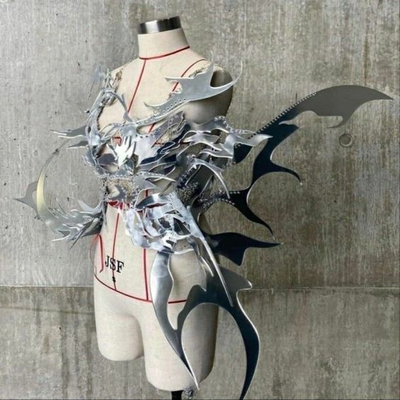 a silver metallic bodice with sharp, angular edges and a winglike design is displayed on a white mannequin against a textured gray wall. the lighting is bright and highlights the intricate details of the piece.