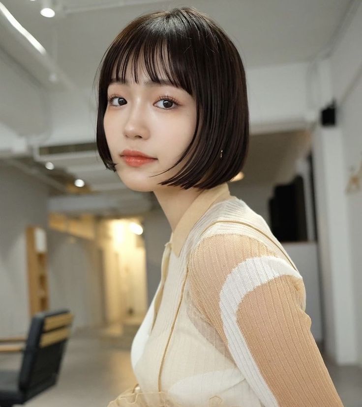 a young asian woman with dark hair styled in a bob with bangs, wears a white and beige striped cardigan and looks at the camera. she is posed in a studio setting with a white wall and light background. she has light makeup and appears to be in her twenties.