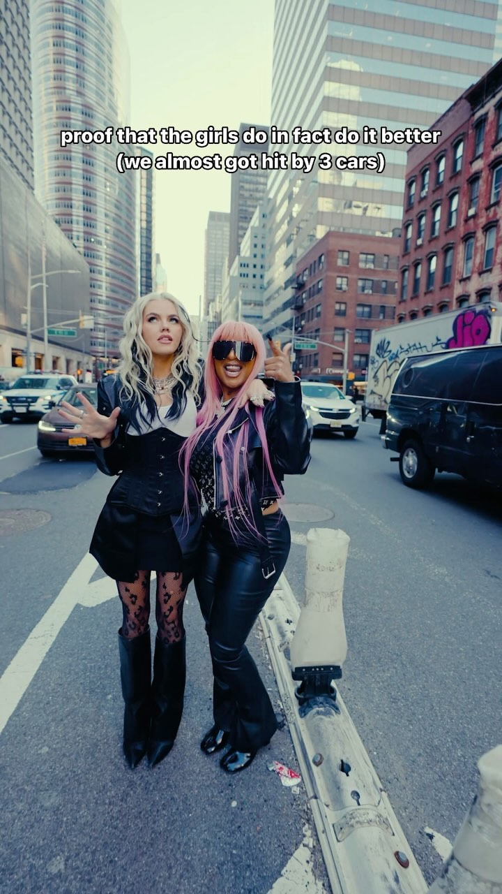 two women pose in the middle of a city street, surrounded by buildings and traffic. one woman has blonde hair and is wearing a black outfit with leggings and boots. the other has pink hair and is wearing sunglasses, a leather jacket, and leather pants. text overlay reads "proof that the girls do in fact do it better (we almost got hit by 3 cars)."