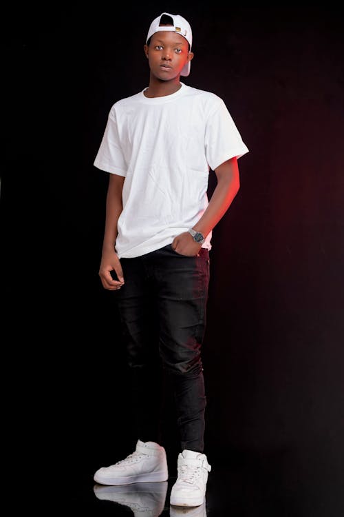 the image shows a young black man standing against a black background. he is wearing a white tshirt, black pants, white sneakers, and a white baseball cap.  he has a serious, confident expression on his face. the lighting is primarily dark, but there is a subtle red glow visible in the background. the overall style is casual and stylish, with a hint of streetwear influence. the photo appears to be a professional studio portrait.