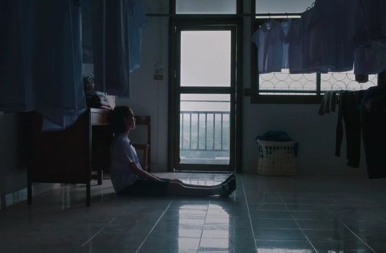 a young woman in a school uniform sits on the floor of a dimly lit room. clothes hang on a rack behind her, and a window and door are visible in the background. the overall atmosphere is one of sadness and contemplation.