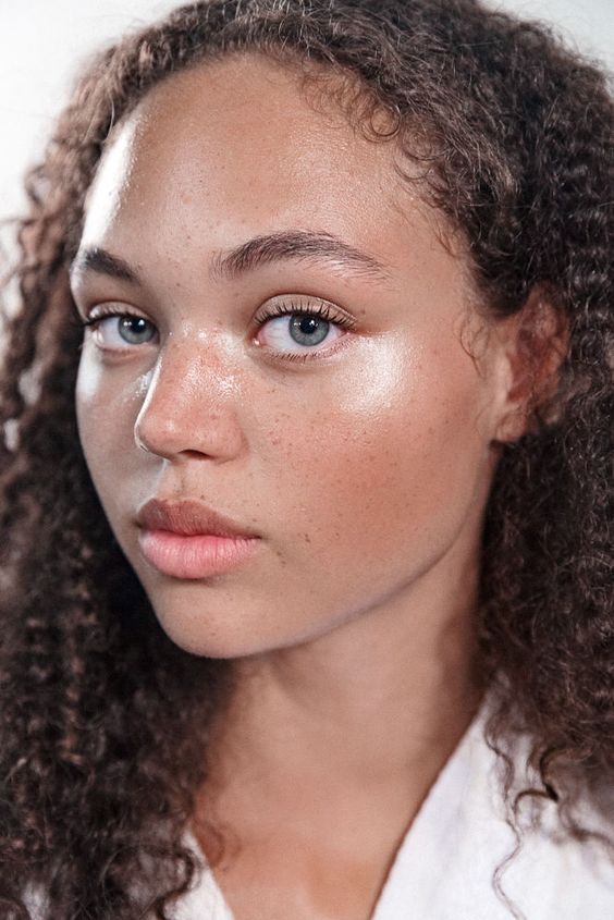 a closeup portrait of a beautiful woman with curly hair, freckles, and blue eyes. she is looking directly at the camera. her skin appears dewy and she seems to be wearing minimal makeup.