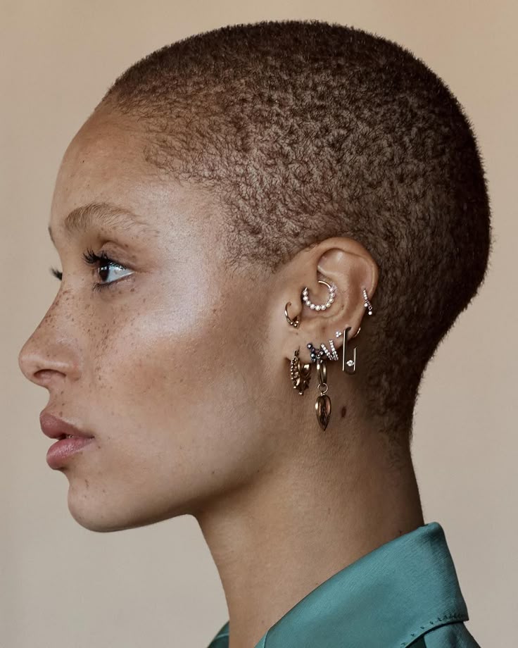a profile shot of a woman with short hair, wearing multiple earrings in her ear. she has freckles and is wearing a green shirt. the focus is on her ear and the various types of earrings she has, including hoops, studs, and pearl earrings.