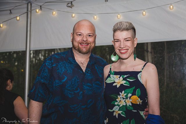 a medium shot shows a bald man and a woman with short, lightcolored hair smiling at the camera. the man has a short, reddish beard and is wearing a buttondown shirt with a blue floral pattern. the woman has fair skin and is wearing a dark blue dress with white floral patterns and citrus fruit. she is also wearing large, light green, round earrings. they are standing close together outdoors under a tent adorned with lights. the man has dark complexion, while the woman has fair complexion. a woman with her hair tied back is visible at the left side, wearing a black top.
