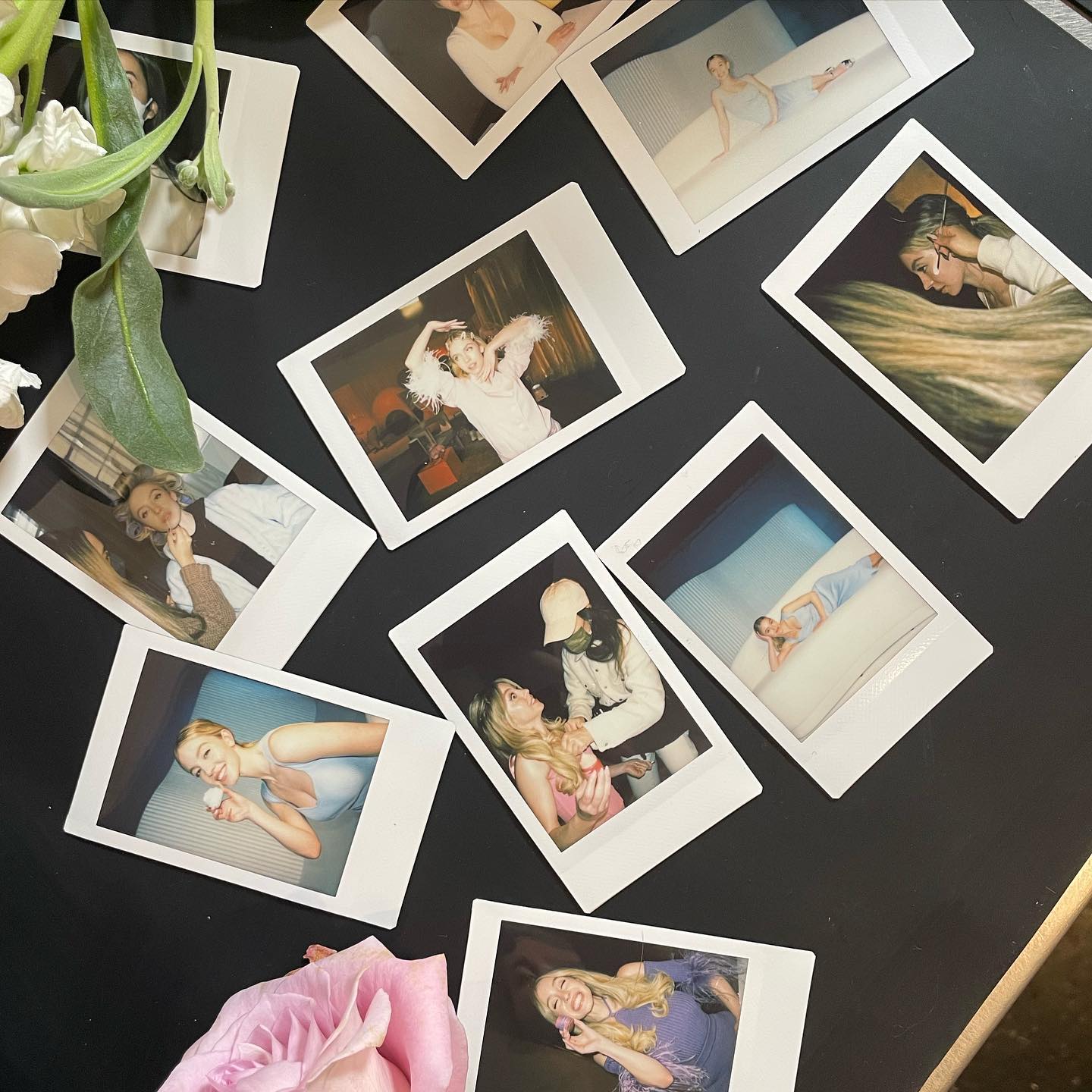a flatlay image showcasing numerous scattered polaroid photos on a dark surface. the photos capture various behindthescenes moments of a photoshoot, including models getting their makeup done, posing, and interacting with the crew.  the photos offer a glimpse into the creative process.  a pink rose is partially visible in the bottom left corner, adding a touch of elegance to the scene. the overall vibe is artistic and messy, reflecting the spontaneous nature of a photoshoot.