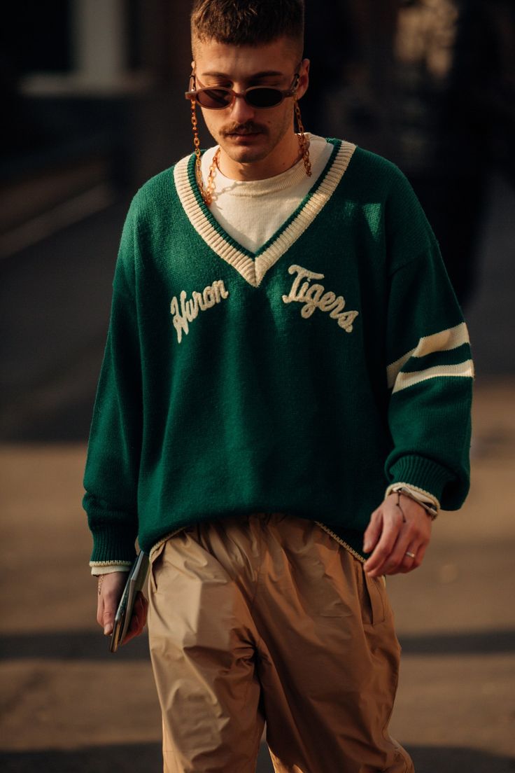 a man wearing a green sweater with white stripes and "wuron tigers" written on it, brown pants, gold chain with sunglasses, looking down at the ground with a casual and trendy street style look.
