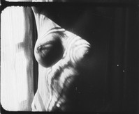 the image is a black and white, closeup film still or photograph. it appears to be a portrait of a woman's upper body, focusing on her breast and shoulder.  the lighting is dramatic, creating strong shadows and highlights.  the background is blurry, suggesting a window with curtains or a sheer fabric. the overall mood is sensual, intimate, and somewhat mysterious due to the darkness and the suggestive nature of the partially visible body. the photographic style is reminiscent of older film or darkroom techniques, with a grainy texture and high contrast. the image's composition is somewhat abstract, with the subject's features softened by the shadows.