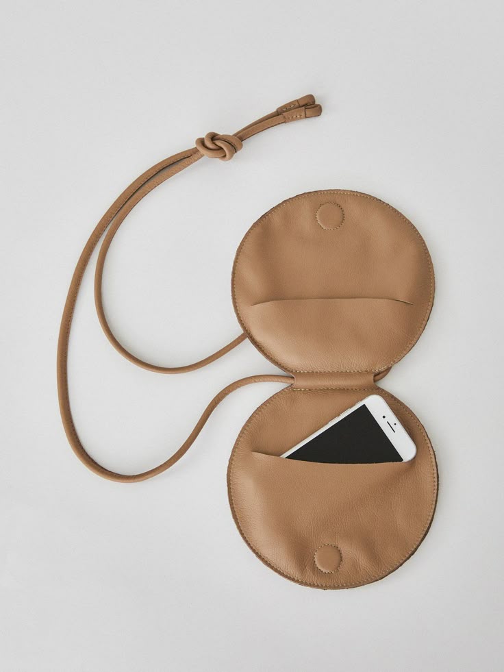 a tancolored leather crossbody bag is shown open with a smartphone inside. the bag is made of two circular pouches with a connecting strap. the strap is knotted near the top, and the smartphone is visible inside the bottom pouch.