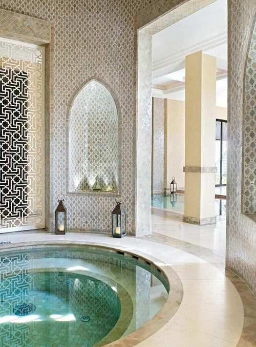 a circular jacuzzi pool sits in a luxurious, intricately tiled bathroom with moroccaninspired arches and geometric patterns. the space is bright, lit by both natural light streaming in from the adjoining pool area and artificial light from lanterns. the ambiance is serene and relaxing, indicative of a spa or highend bathroom setting.
