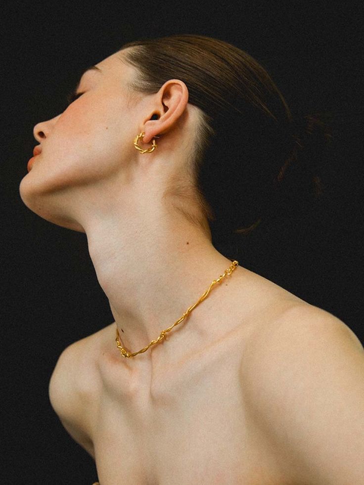 the image is a closeup portrait of a woman's face and upper body, showcasing gold jewelry against a stark black background.  she is wearing a delicate, twisted gold necklace and matching twisted gold hoop earrings. the woman's pose and the minimalist style of the jewelry create a sophisticated and elegant look. the focus is clearly on the jewelry and the model’s skin texture and features. the overall mood is serene and fashionoriented.