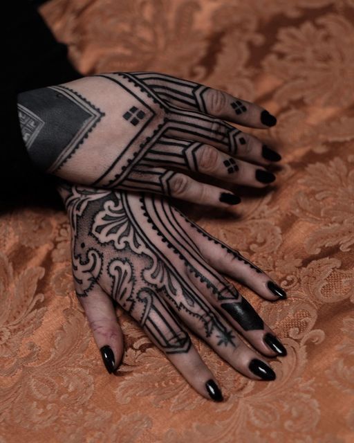 the image shows two hands with intricate black ink tattoos. one hand has a solid black square on the back of the hand with geometric patterns extending to the fingers. the second hand features ornate, hennainspired designs with swirls and detailed line work. the fingernails are painted with black nail polish. the hands are resting on an orange patterned surface. the tattoos cover most of the hands and fingers.