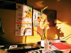 a cozy workspace is bathed in the warm glow of the setting sun. the golden light illuminates a collection of art prints and posters on the wall, casting long shadows across the desk. a vase holding a tall, elegant flower adds a touch of nature to the scene. a laptop, open and ready for work, sits alongside a scattering of papers and art supplies, hinting at creativity and productivity.