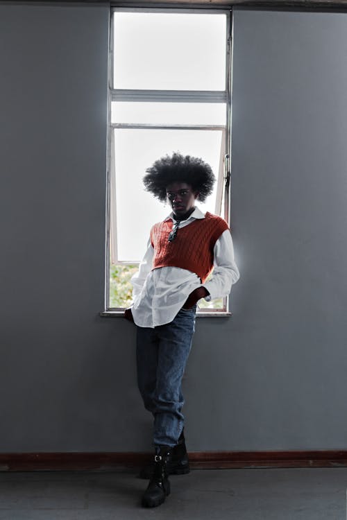 a full shot of a young man with an afro hairstyle standing by a window in a room with grey walls. he's wearing a white shirt over an orange sweater vest, blue jeans, and black boots. his pose is casual, leaning slightly against the window frame with one hand in his pocket.  the overall mood is contemplative and slightly moody. the image has a stylish and somewhat editorial feel.
