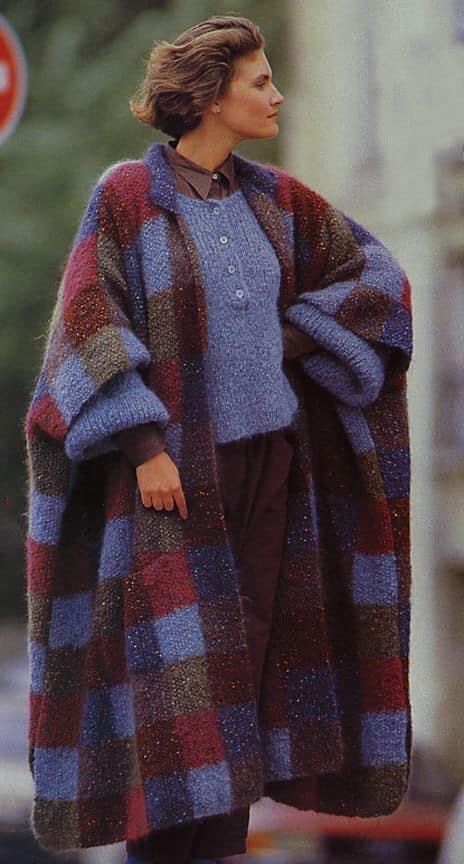 a woman is wearing a blue sweater and a large, colorful, checkered knit coat.