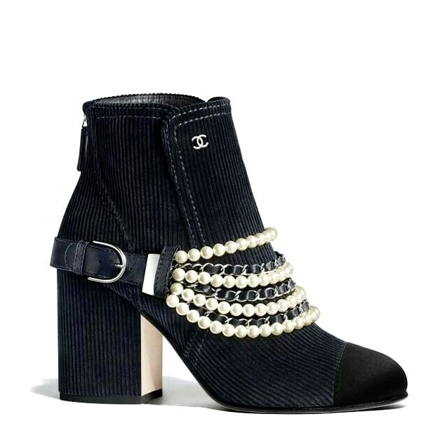 a black corduroy chanel ankle boot with a block heel, a cap toe, a leather strap with a buckle around the ankle, and strands of pearls and a silver chain across the vamp is centered in the image against a white background. the heel has a creamcolored accent piece. the boot has a zipper on the back and a silver chanel logo near the top edge.