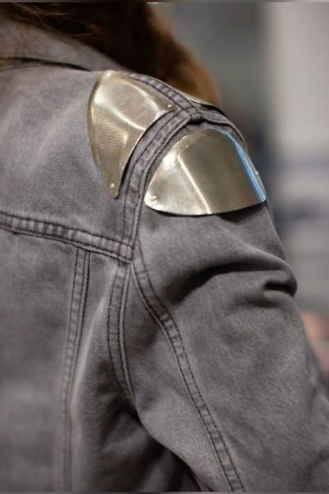 a closeup of a person wearing a gray denim jacket with silver metal shoulder pads. the shoulder pads are shaped like triangles and are attached to the jacket with stitching. the jacket is textured and has a stylish, edgy look.
