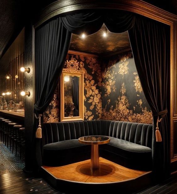 a dark, intimate booth with a velvet sofa and a round table in the center. the walls are covered in gold wallpaper and there are ornate lamps and mirrors hanging from the ceiling. the booth is situated in a corner, with black velvet curtains hanging from the ceiling and framing the seating area. the wood floor is polished and gleaming.