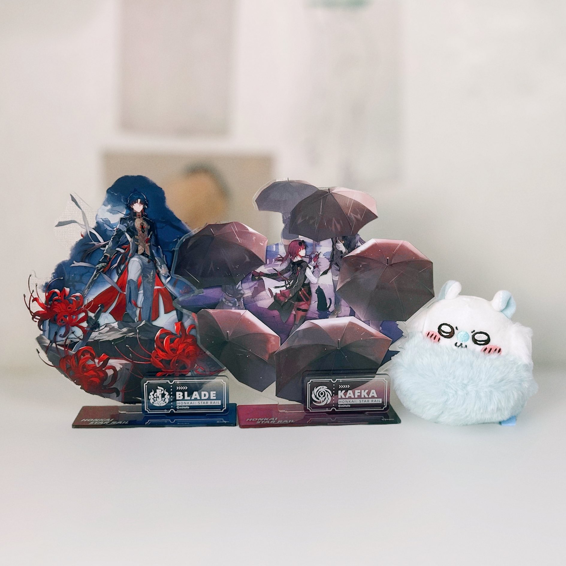 an image of honkai star rail merchandise, showcasing figures of the characters blade and kafka, displayed on their respective acrylic stands. there's a cute plush toy resembling a white character from the game placed on the table. the background is blurred, highlighting the merch.
