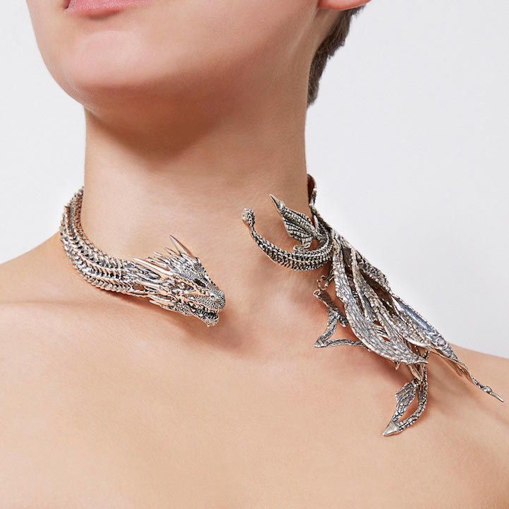 a closeup shot showcases a woman wearing an elaborate silver dragon necklace. the necklace consists of a dragon head on one side of her neck and a winglike structure on the other. the jewelry is highly detailed, with intricate scales and featherlike textures, appearing to be a statement piece of contemporary fashion.