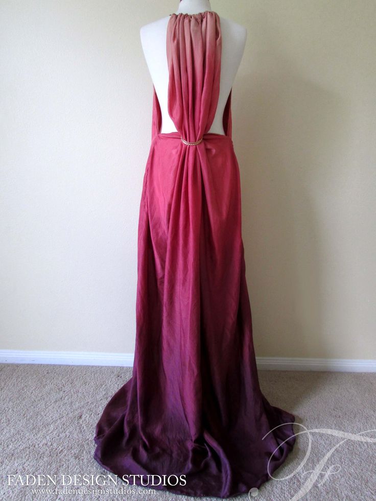 a back view of a long, ombrecolored dress draped on a white mannequin. the dress transitions from a light pink at the top to a deep plum at the bottom. the fabric is gathered at the top of the back and also at the middle where it is held together by a gold ring. the dress is backless and the fabric has a slight train at the bottom. the background consists of a beige wall and a carpeted floor. the words "faden design studios" and their website address are visible at the bottom of the image.